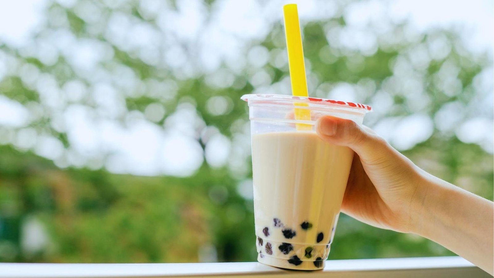 Hand holds sealed milk tea with black tapioca pearls and bright yellow straw outdoors, with blurred green trees in background.