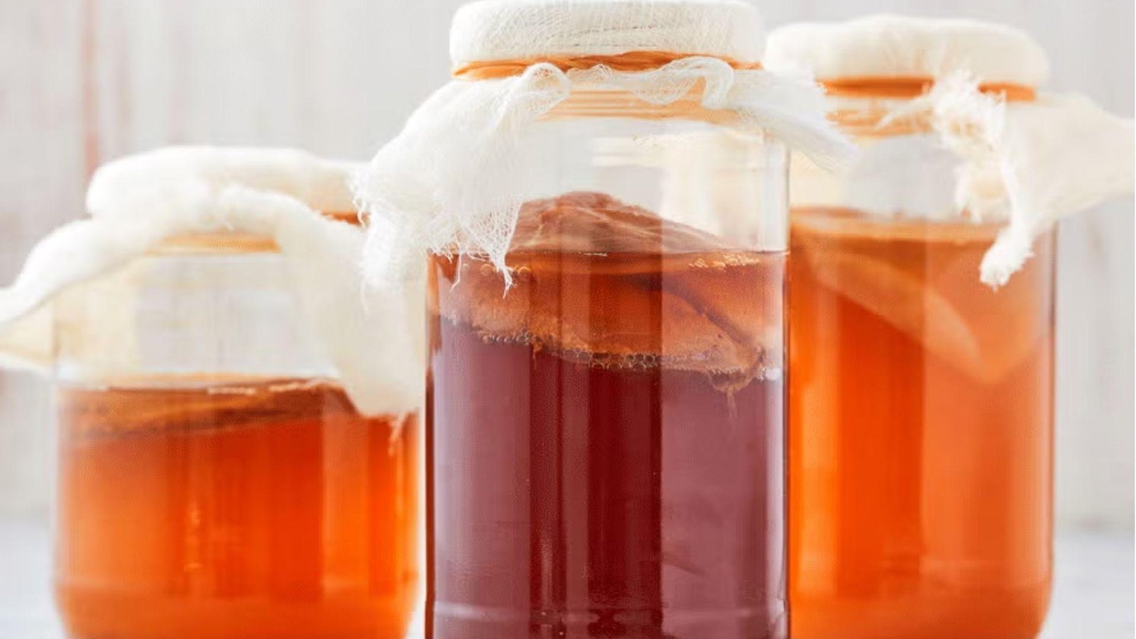 Three glass jars of fermenting kombucha covered with cheesecloth in amber and deep red shades on a white surface.