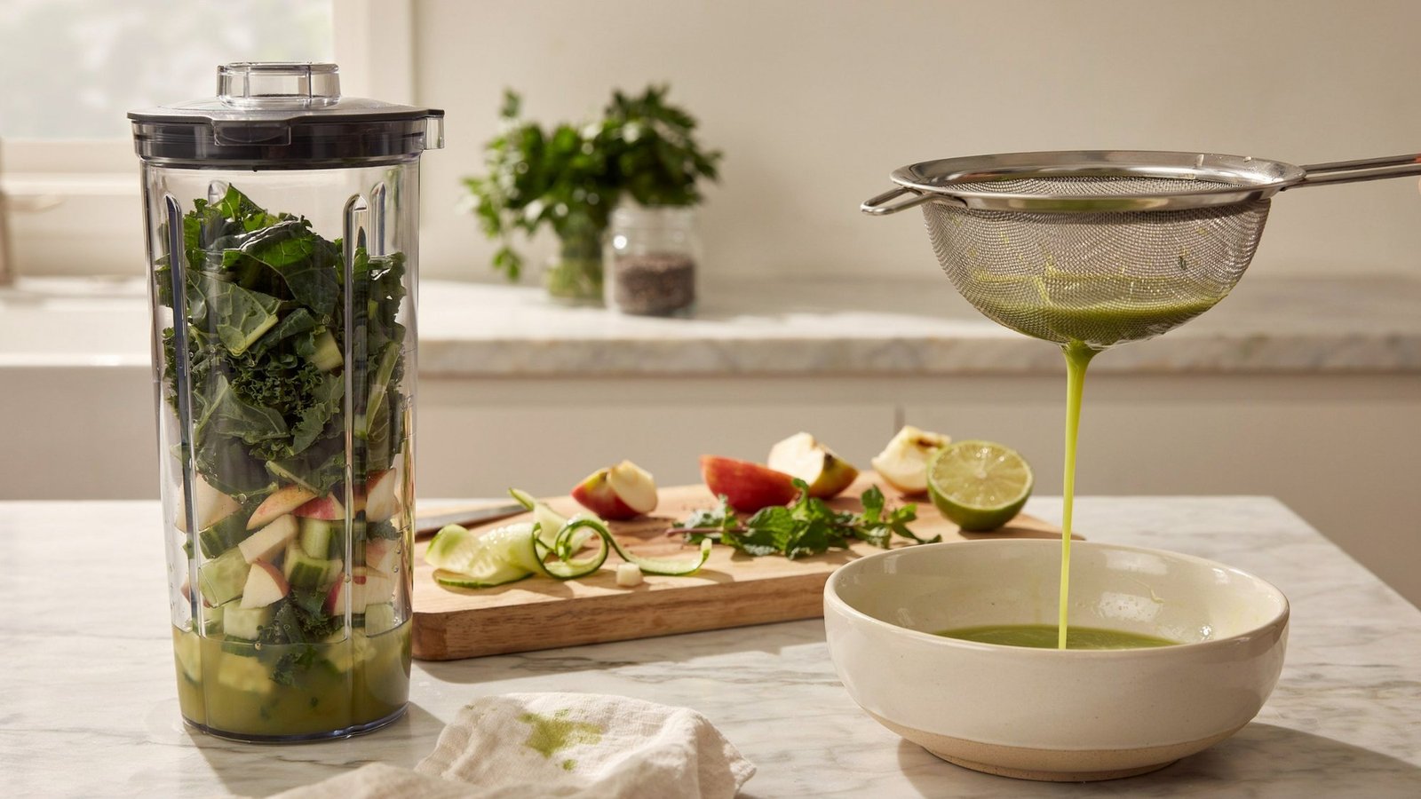 Blender preparing green juice with fruits vegetables and water then strained through mesh to remove pulp in kitchen setup.