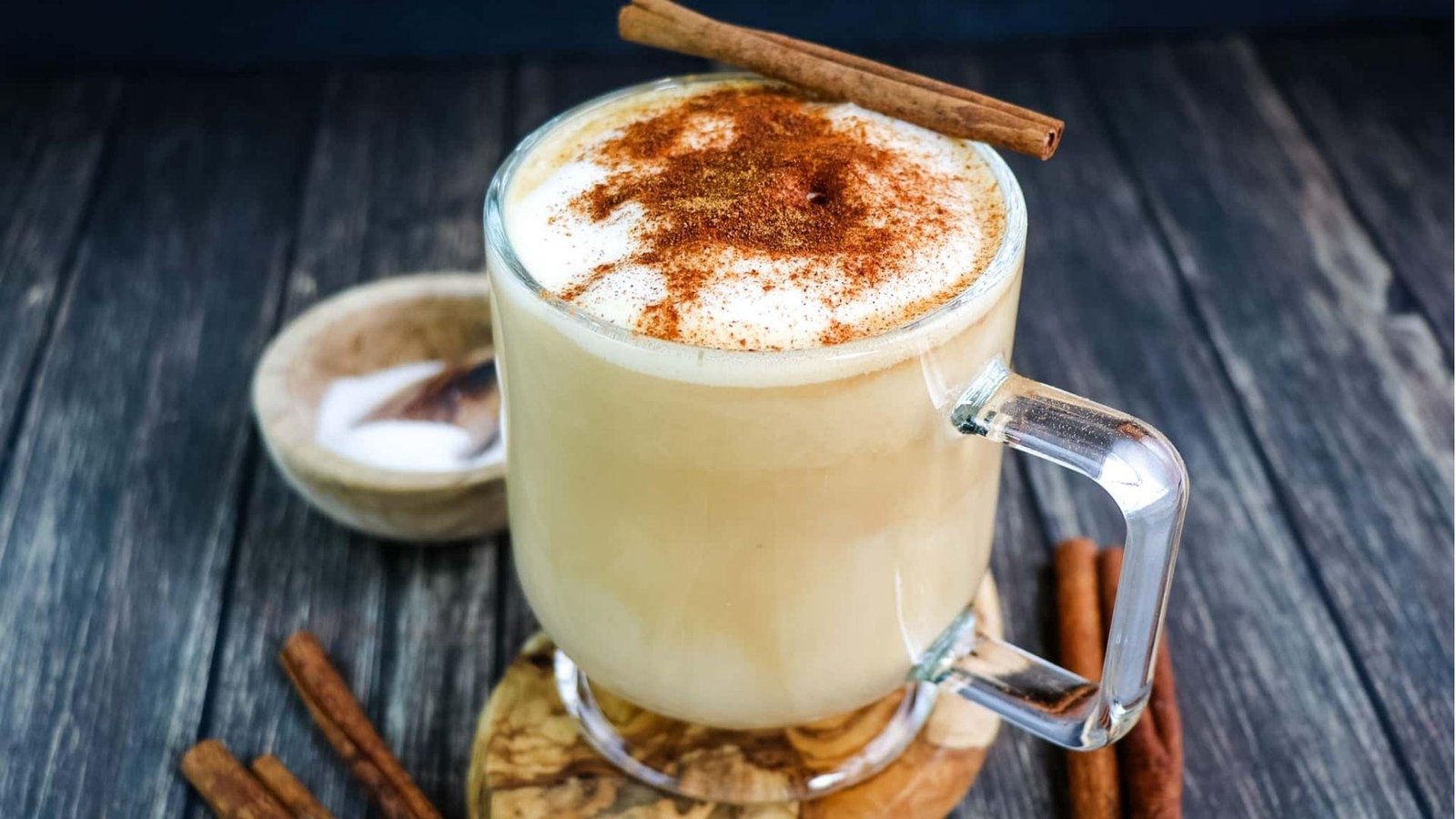 A glass mug of creamy dolce latte topped with thick foam, cinnamon dusting, and a cinnamon stick on a wooden coaster.