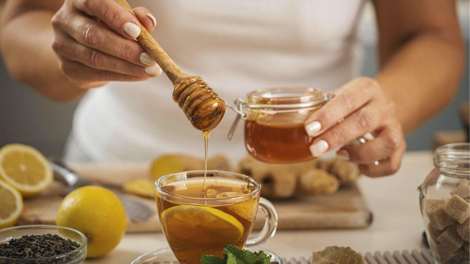A woman pours honey from a jar into lemon tea with ginger, mint, and sugar on the kitchen table.