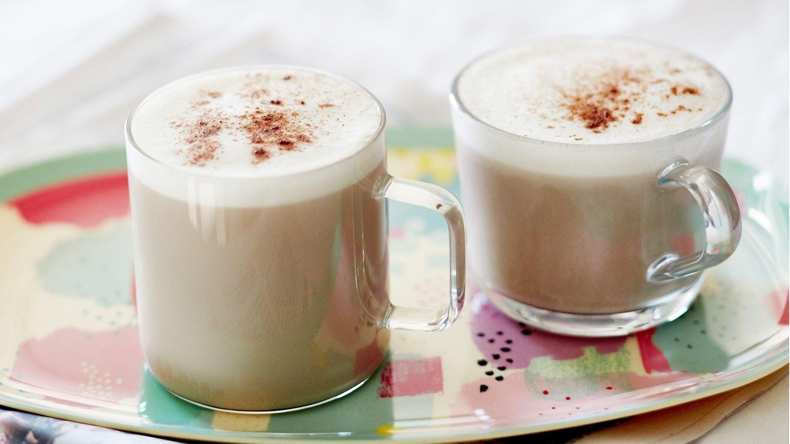 Two glass mugs of creamy chai latte dusted with cinnamon on a colorful decorative tray against a white background.