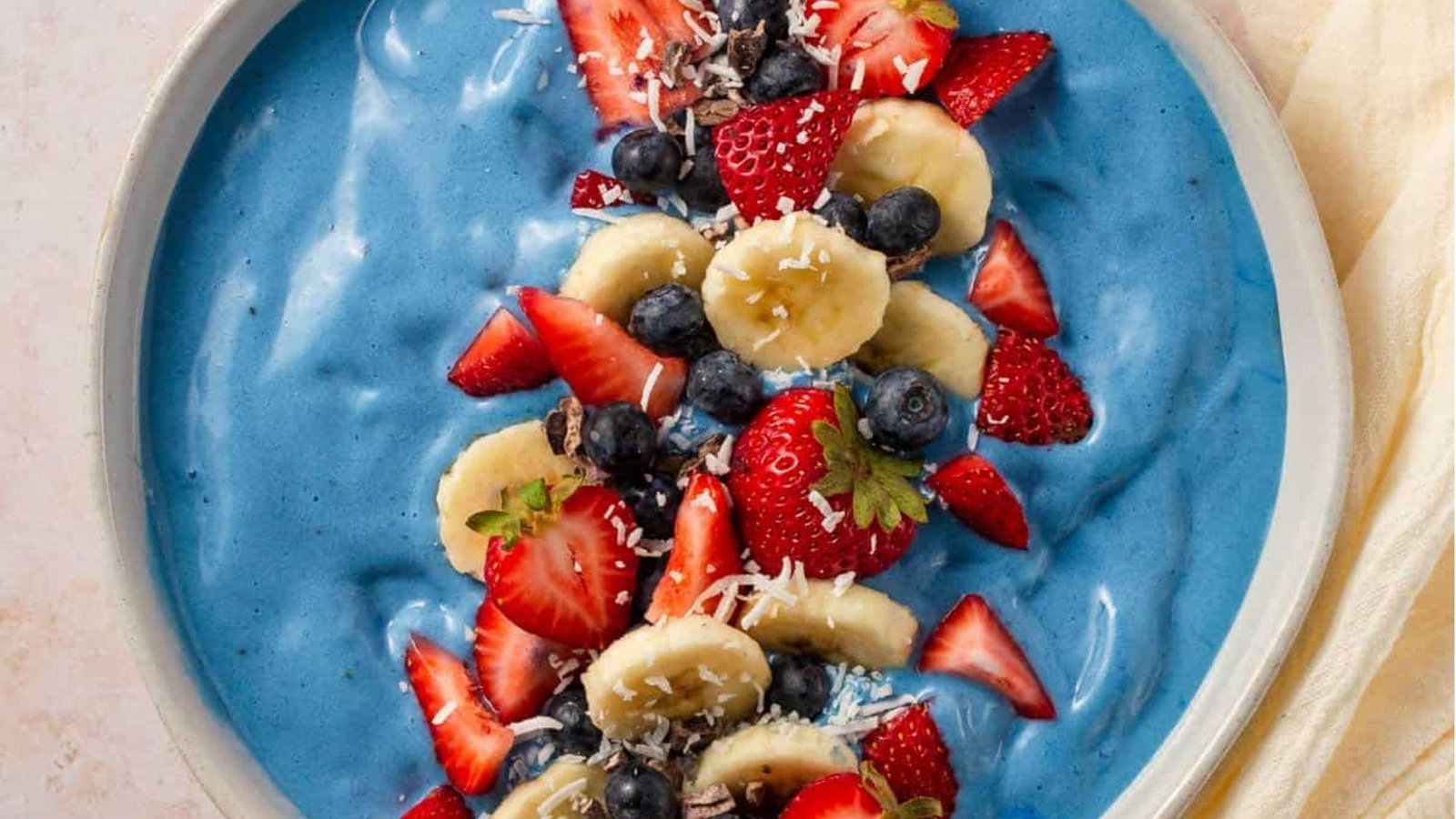 Overhead view of a blue spirulina smoothie bowl topped with a row of strawberries, banana slices, blueberries, and shredded coconut.