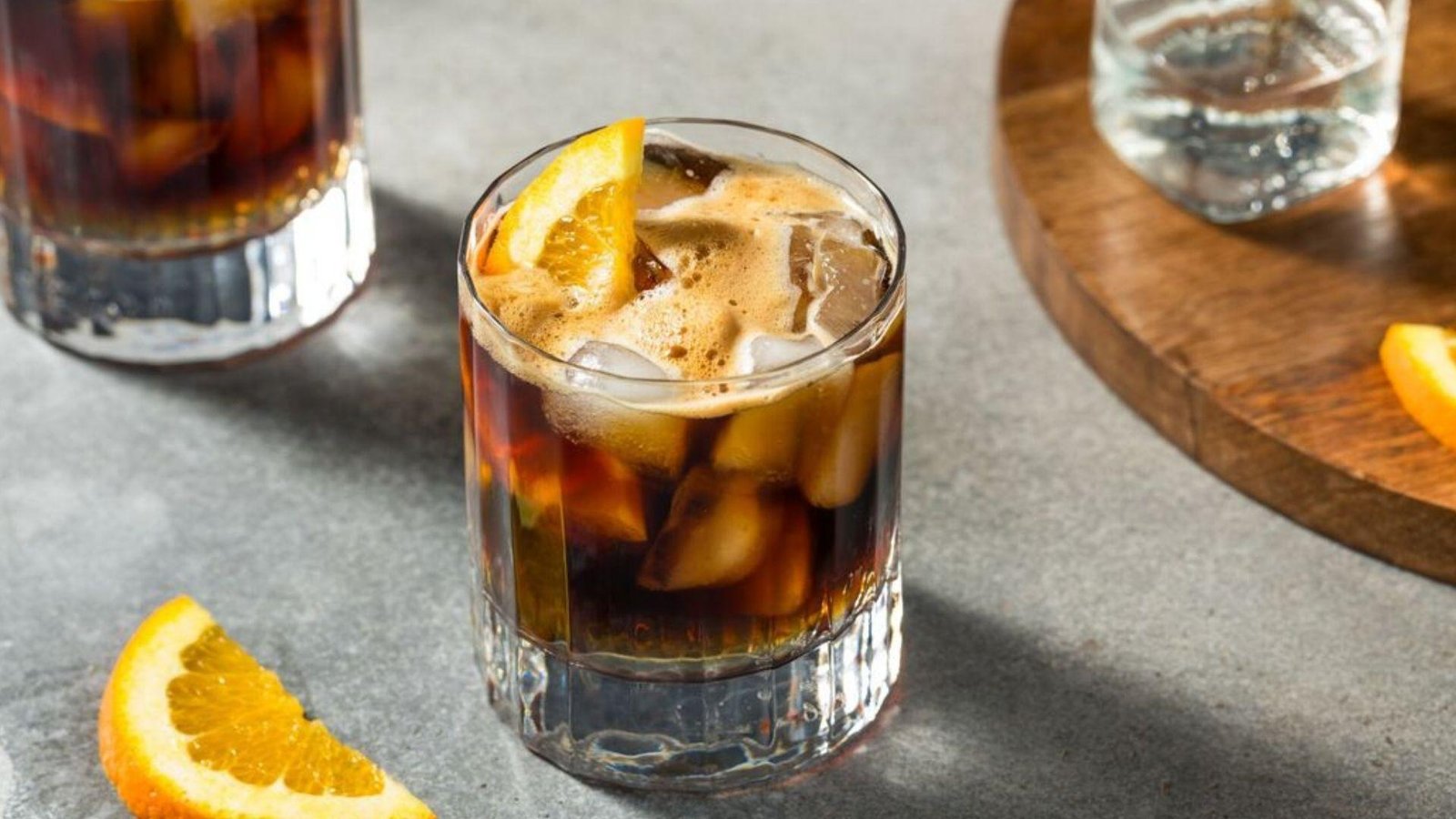 A rocks glass of dark iced coffee garnished with an orange wedge, with coffee foam on top, on a grey surface.