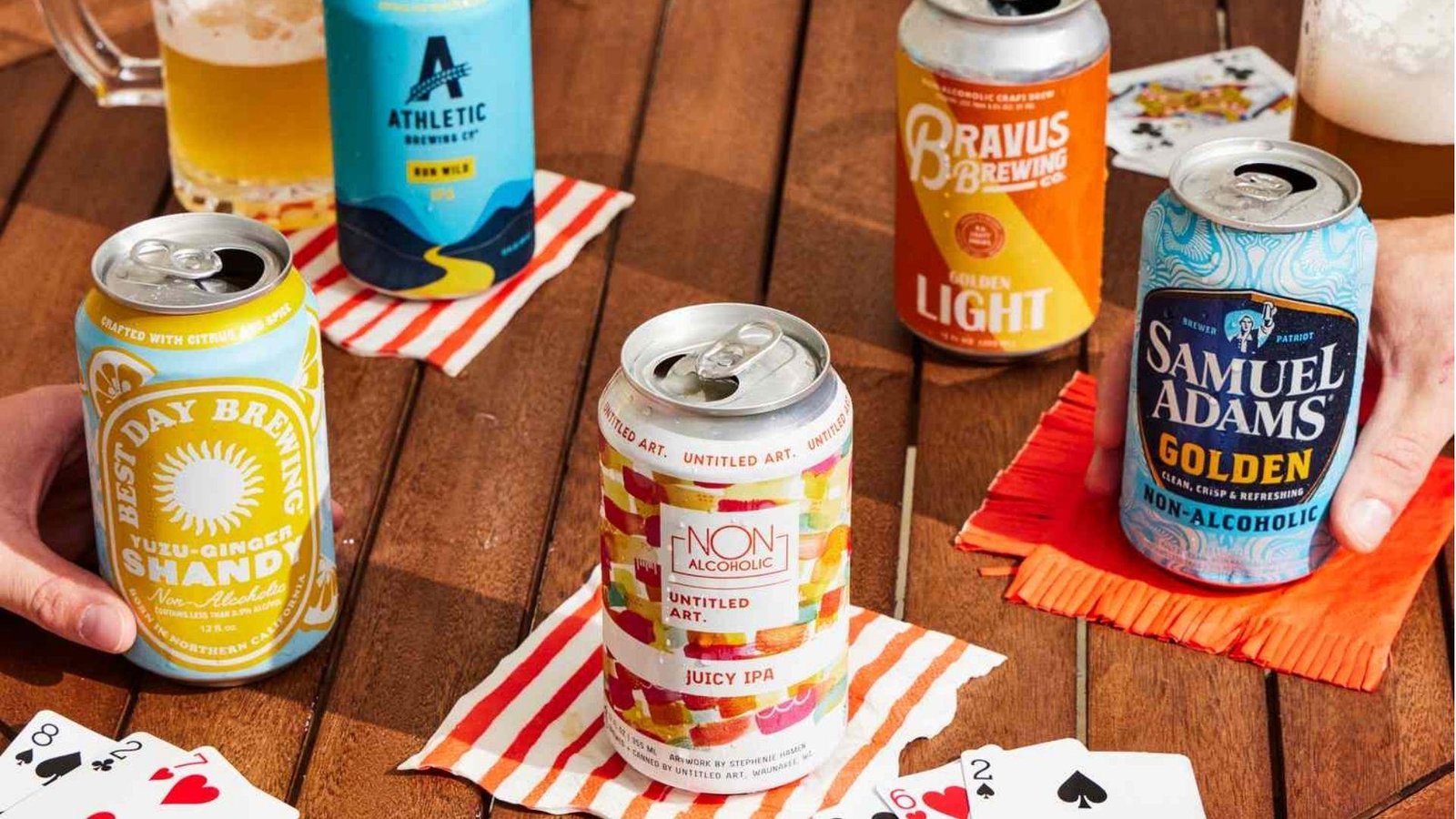Five non-alcoholic beer cans from Best Day Brewing, Athletic Brewing, Untitled Art, Bravus, and Samuel Adams on a wooden table.