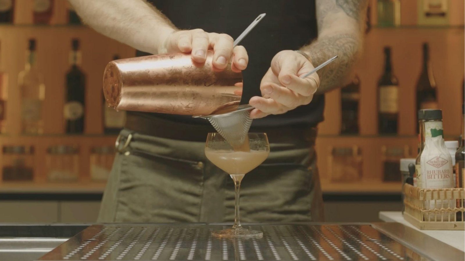 Hands straining a freshly shaken Mezcal Espresso Martini from a copper shaker into a chilled Nick & Nora glass.