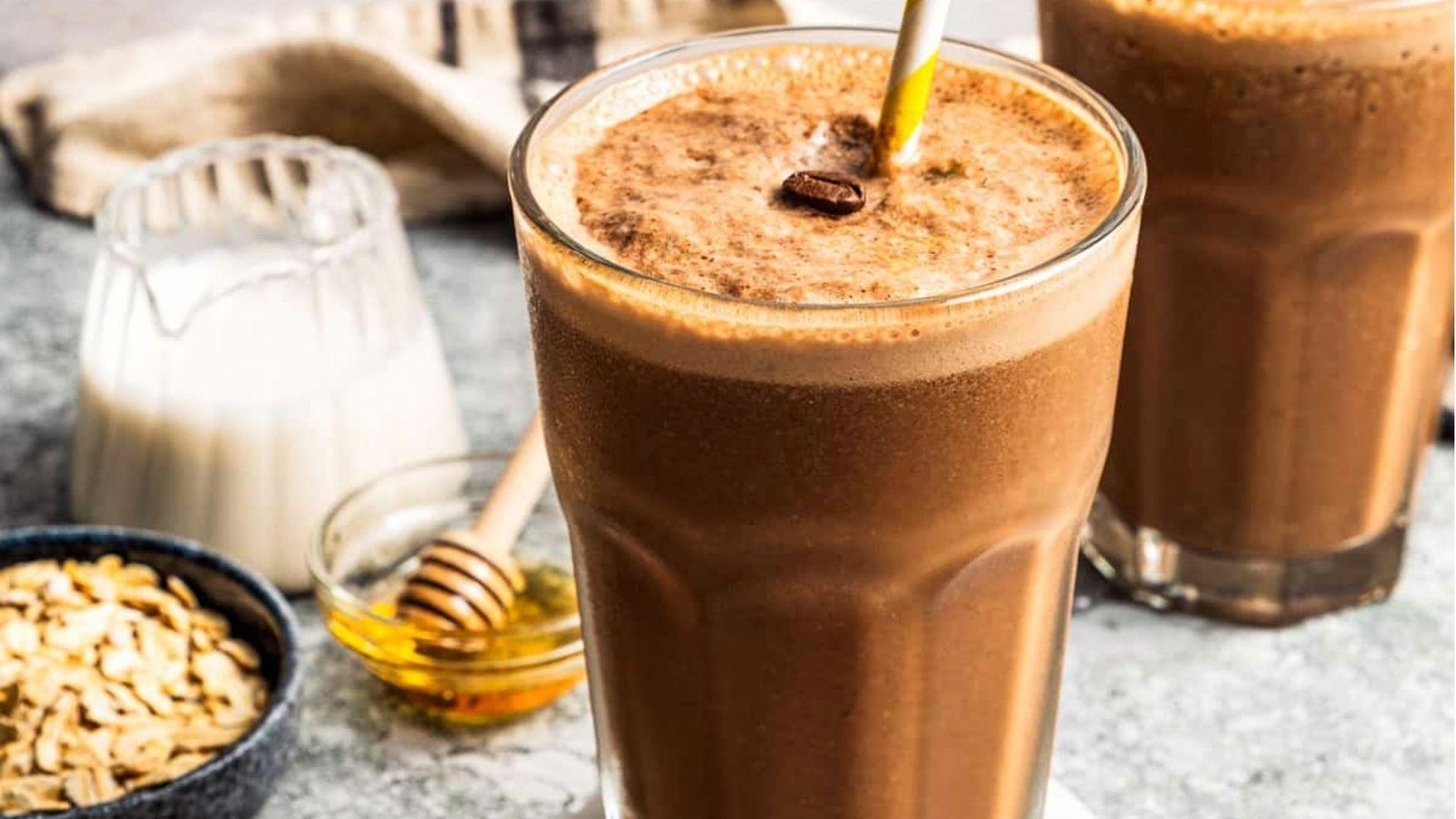 Two tall glasses of chocolate coffee smoothie topped with cinnamon and a coffee bean, with oats, milk, and honey alongside.