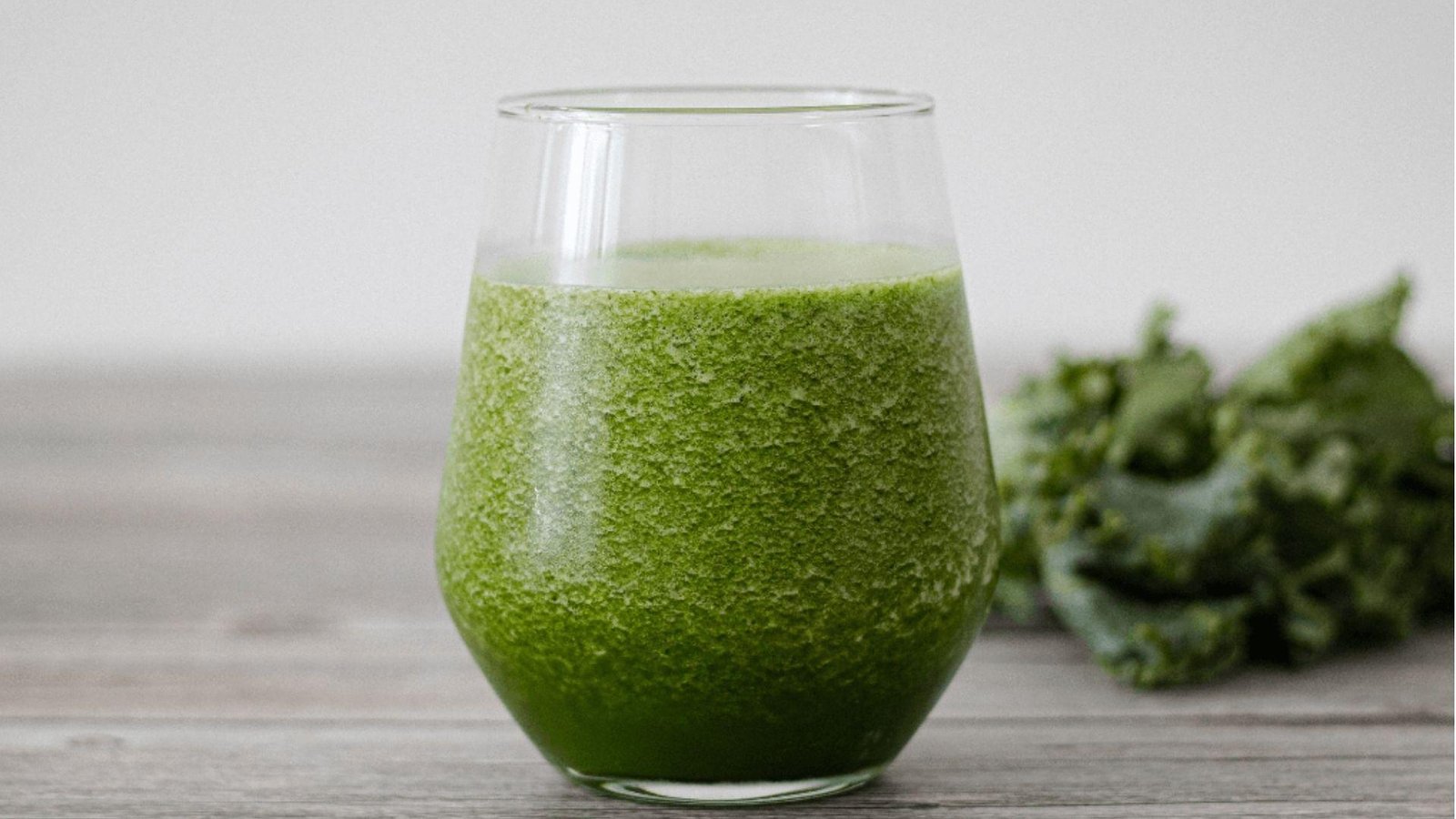 A stemless glass of thick green kale smoothie on a grey wooden surface with fresh kale leaves in the background.