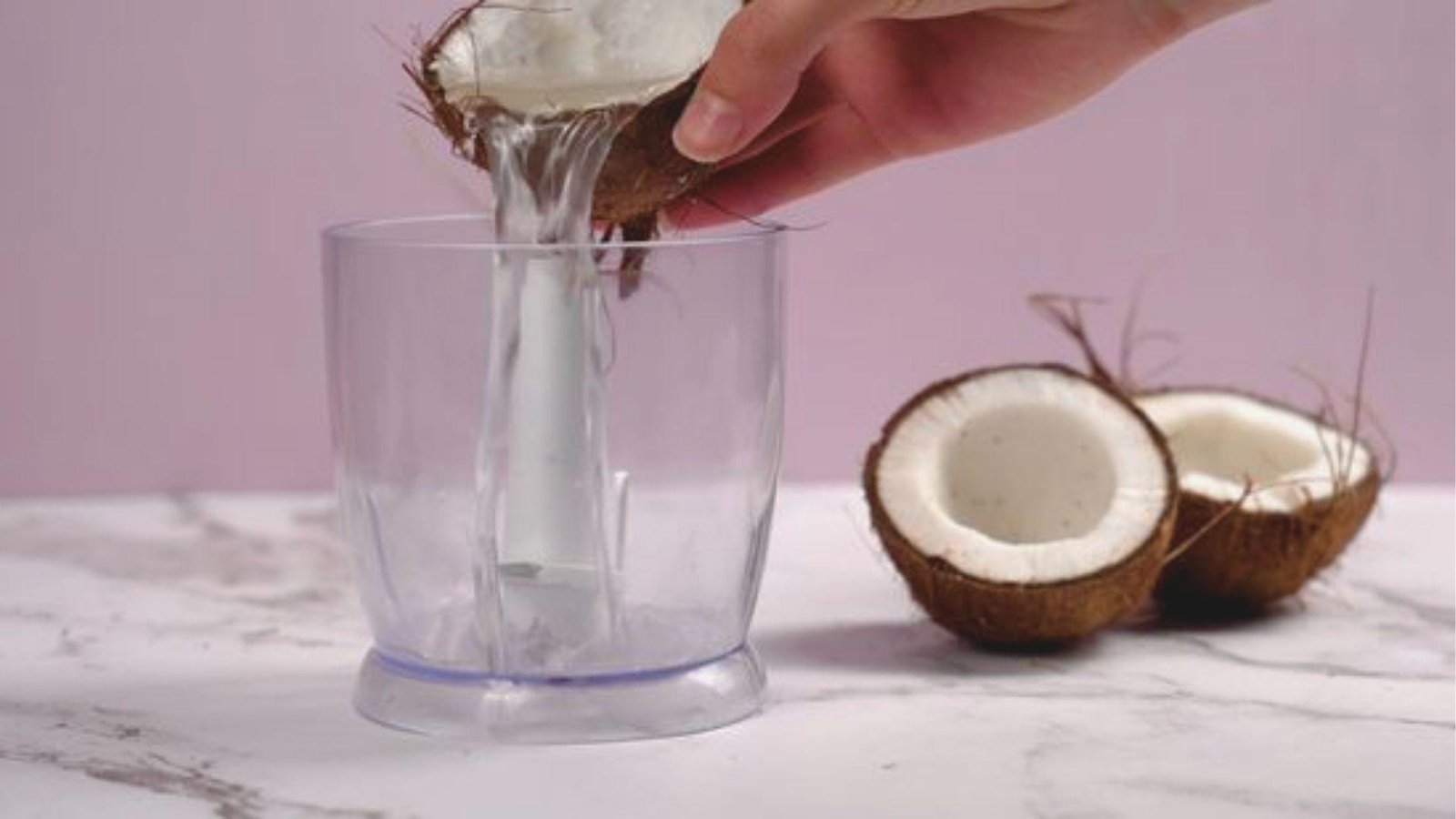 Hand pouring fresh coconut water from a cracked coconut shell into a clear blender cup.
