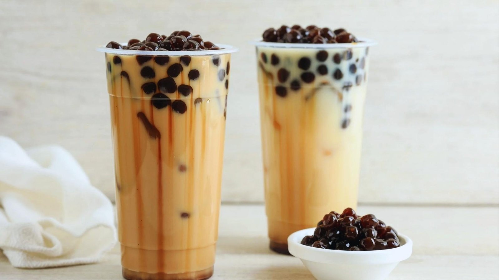 Two tall cups of brown milk tea overflow with glossy black pearls beside a small white bowl of tapioca.