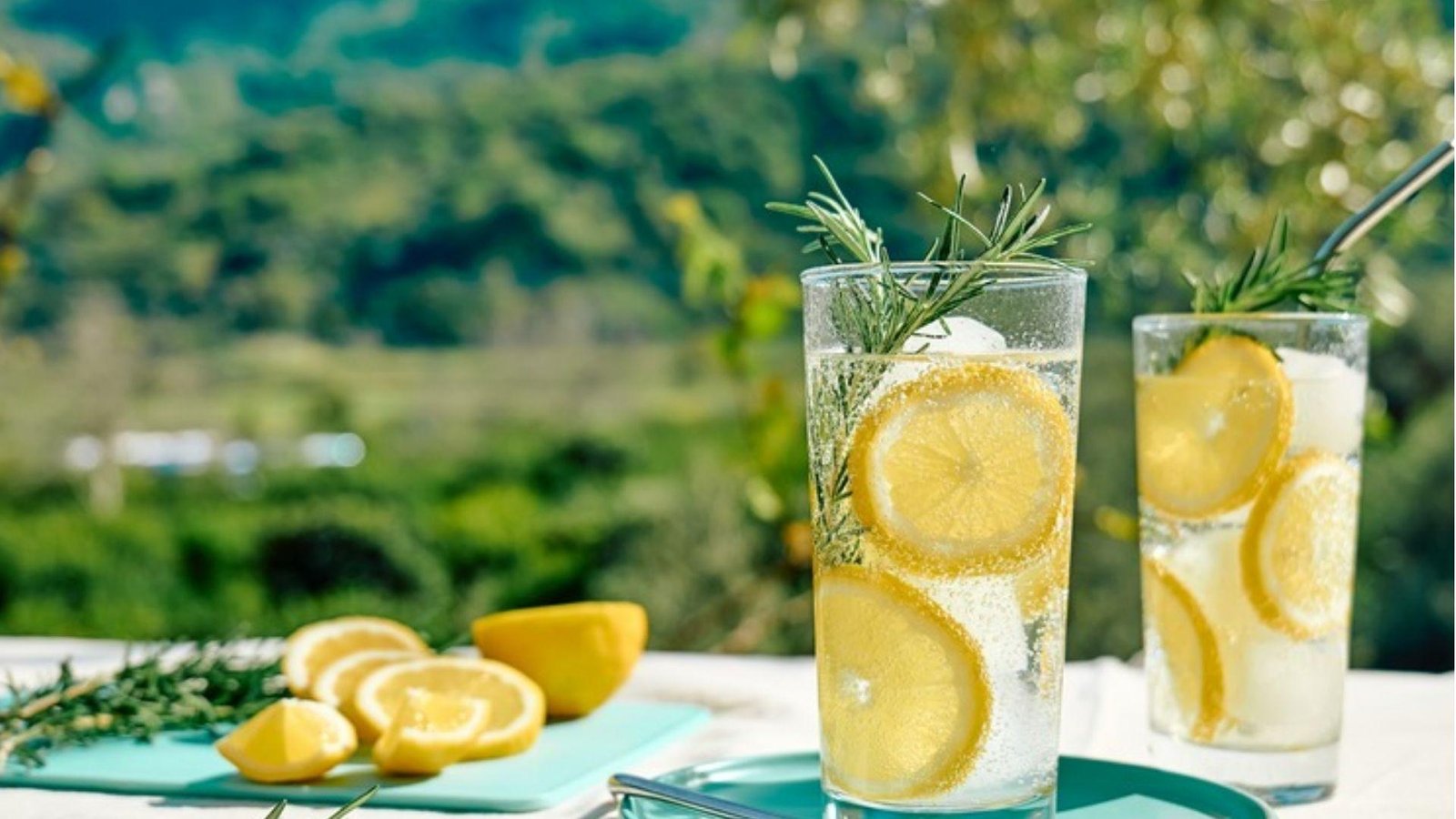Two tall glasses of sparkling lemonade with lemon slices and rosemary sprigs on a white outdoor table with greenery.