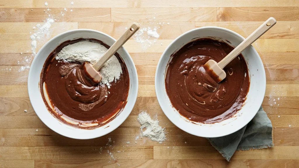 Two white bowls side by side showing brownie batter before and after flour is folded in, on a wooden surface.