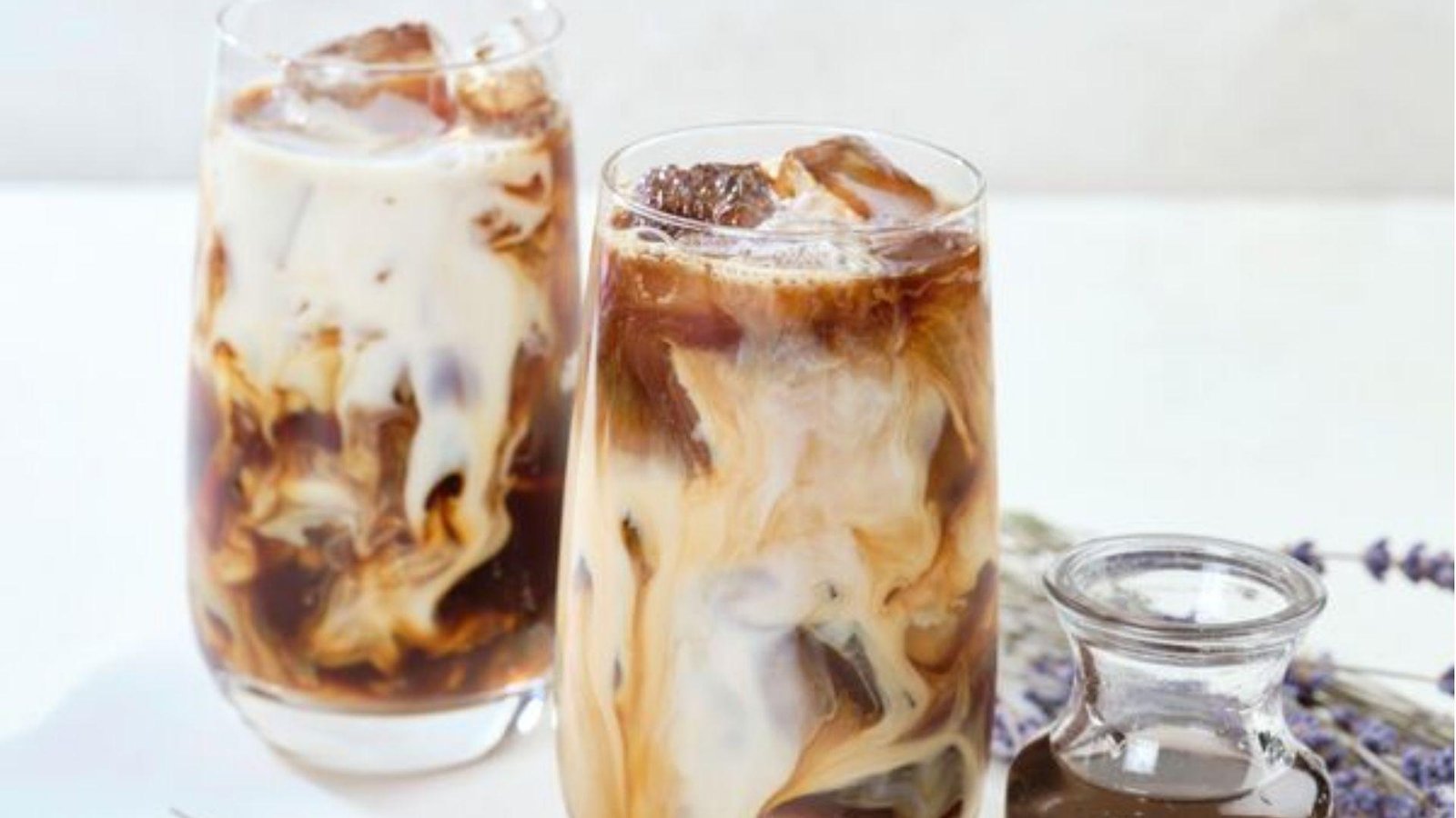 Two tall glasses of iced brown sugar latte brew with swirling cream patterns, beside dried lavender and a syrup jar.