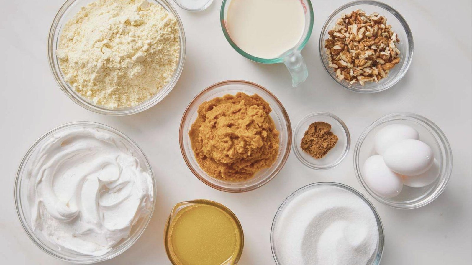 Flat lay of Pumpkin Crunch Recipe ingredients in glass bowls: cake mix, pumpkin puree, eggs, sugar, spices, chopped pecans, and milk.