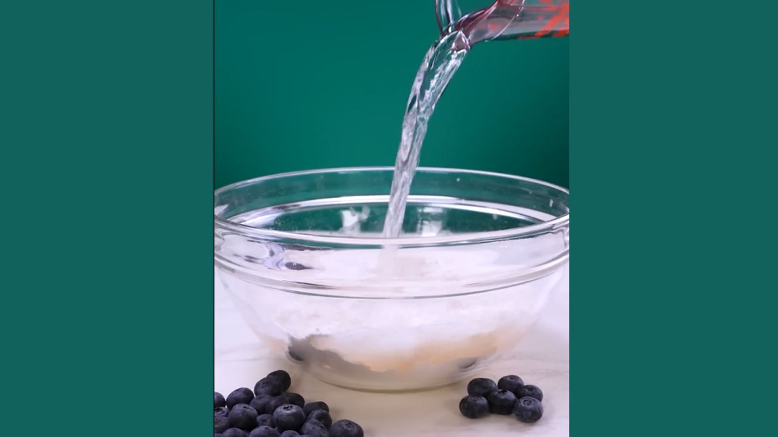 Water poured from measuring cup into glass bowl with flour and blueberries on counter for blue waffle batter