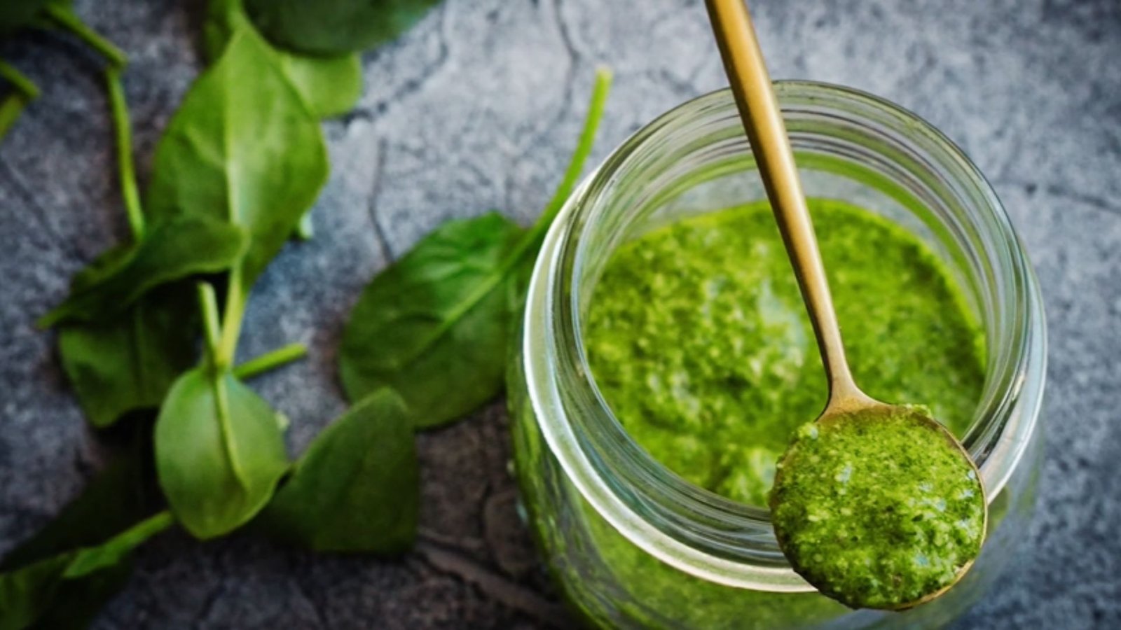 Gold spoon scooping fresh homemade pumpkin seed pesto from jar with spinach leaves nearby