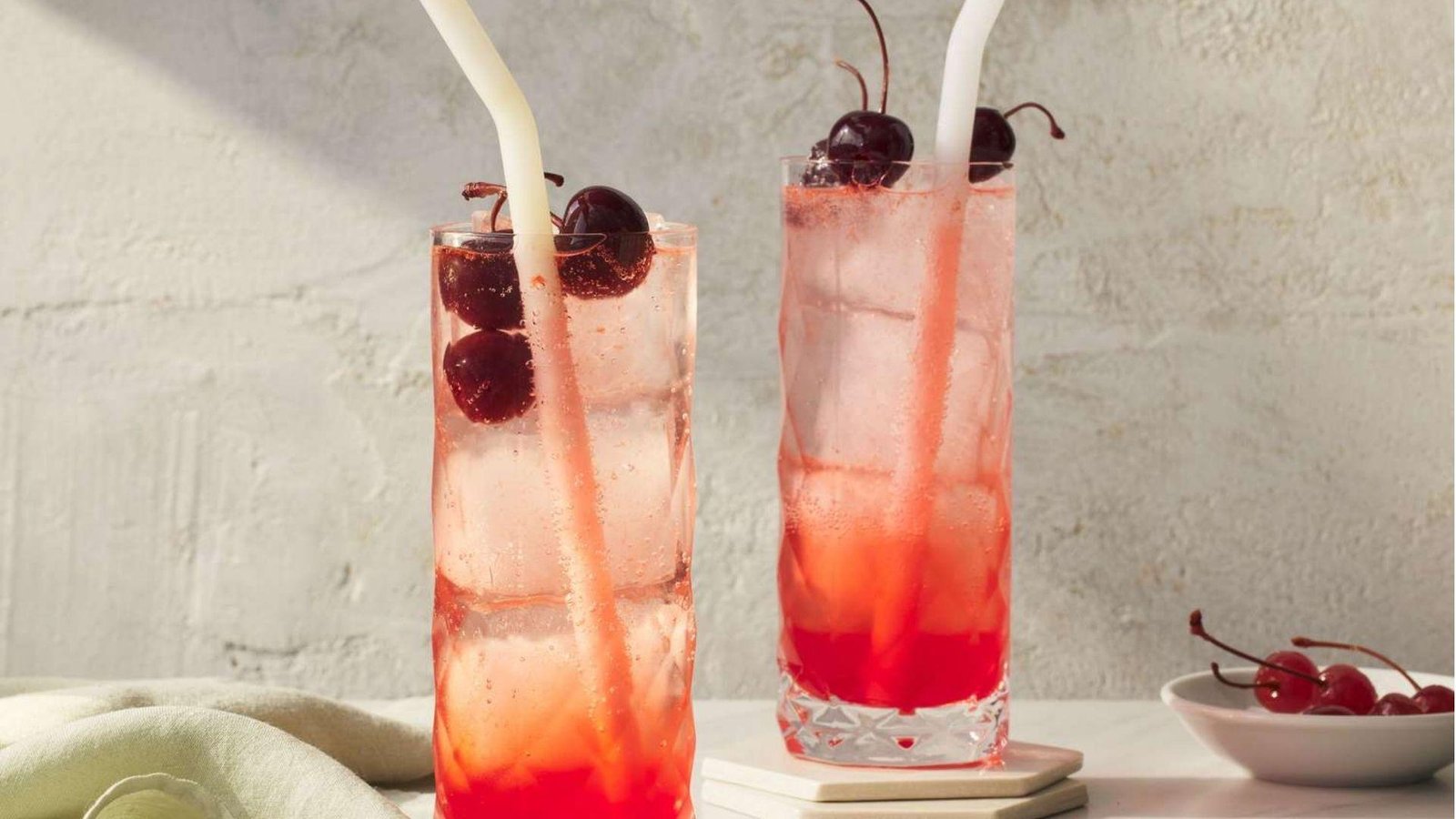 Two tall glasses of shirley temple with ice, dark cherries, and white straws on a light surface beside a cherry bowl.