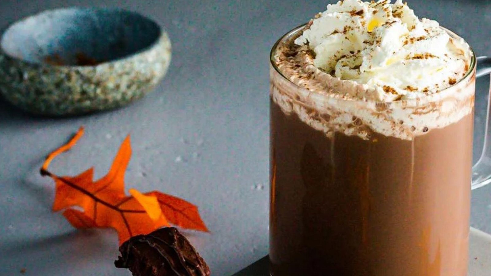 A glass mug of hot chocolate mocha topped with whipped cream and pumpkin powder, with an autumn leaf and chocolate beside it.