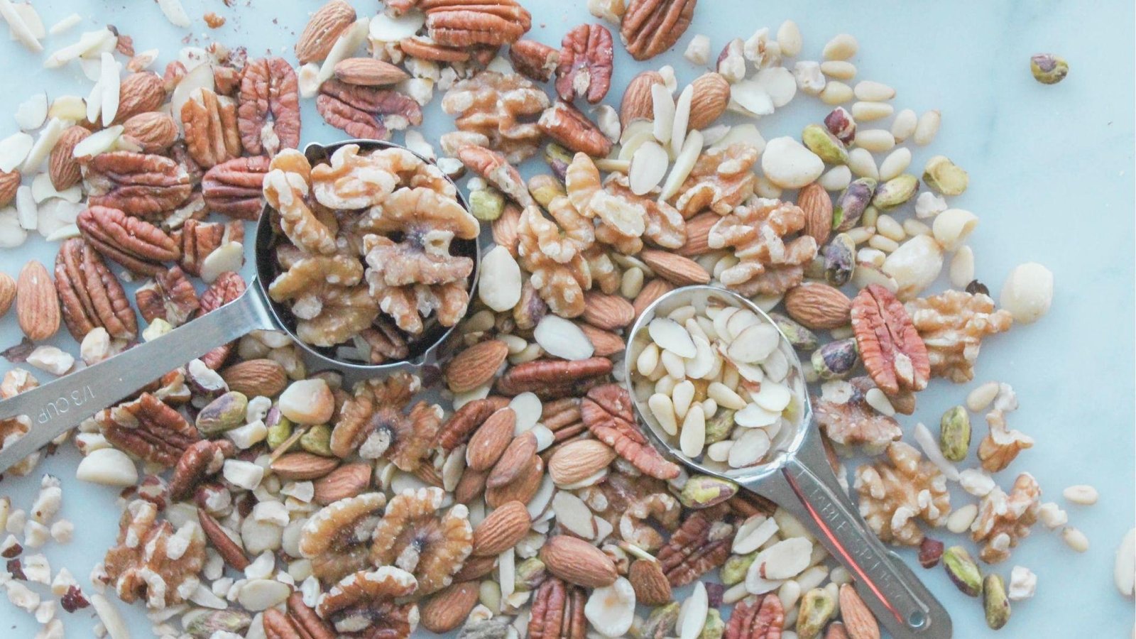 Overhead view of assorted chopped nuts including walnuts, pecans, almonds, and pistachios scattered on a light surface.