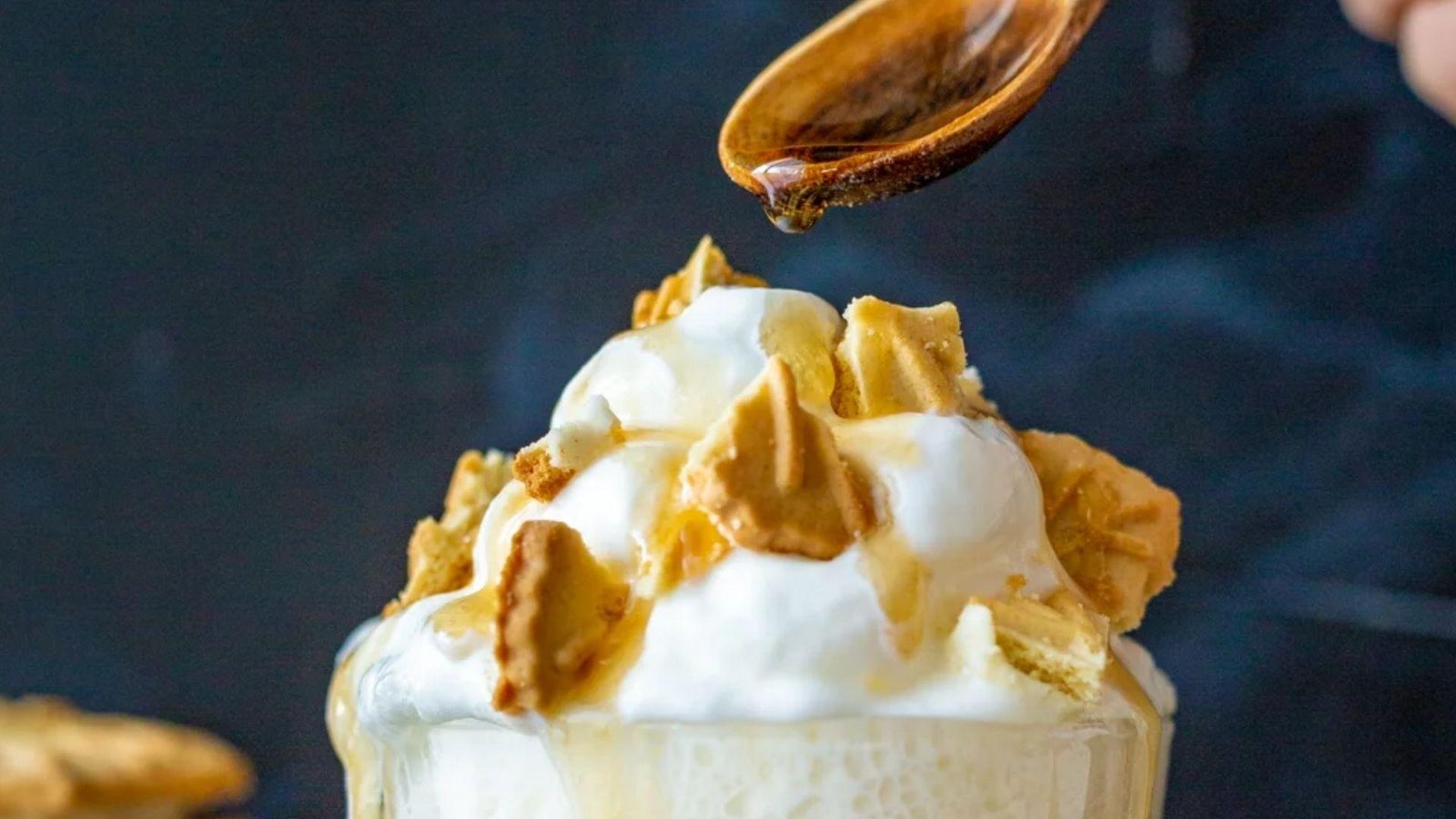 A close-up of a whipped cream latte topped with golden cookie pieces and maple syrup being drizzled from a wooden spoon against a dark blue background