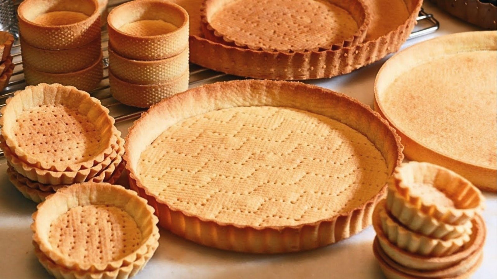Golden baked pate sablee tart shells and mini tartlets cooling on rack, ready to fill.