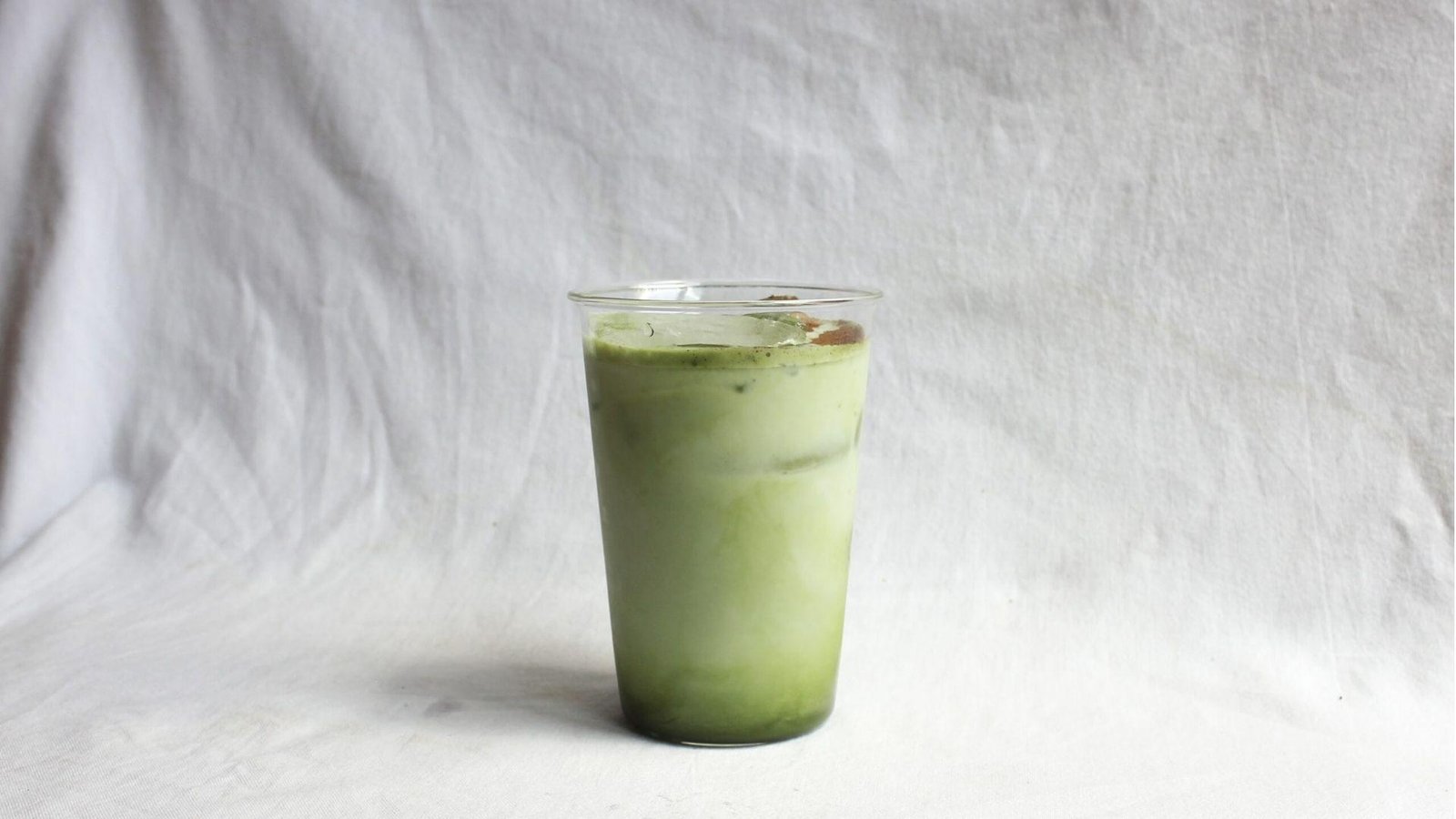 A simple clear plastic cup of iced matcha green tea latte against a plain white linen fabric background.