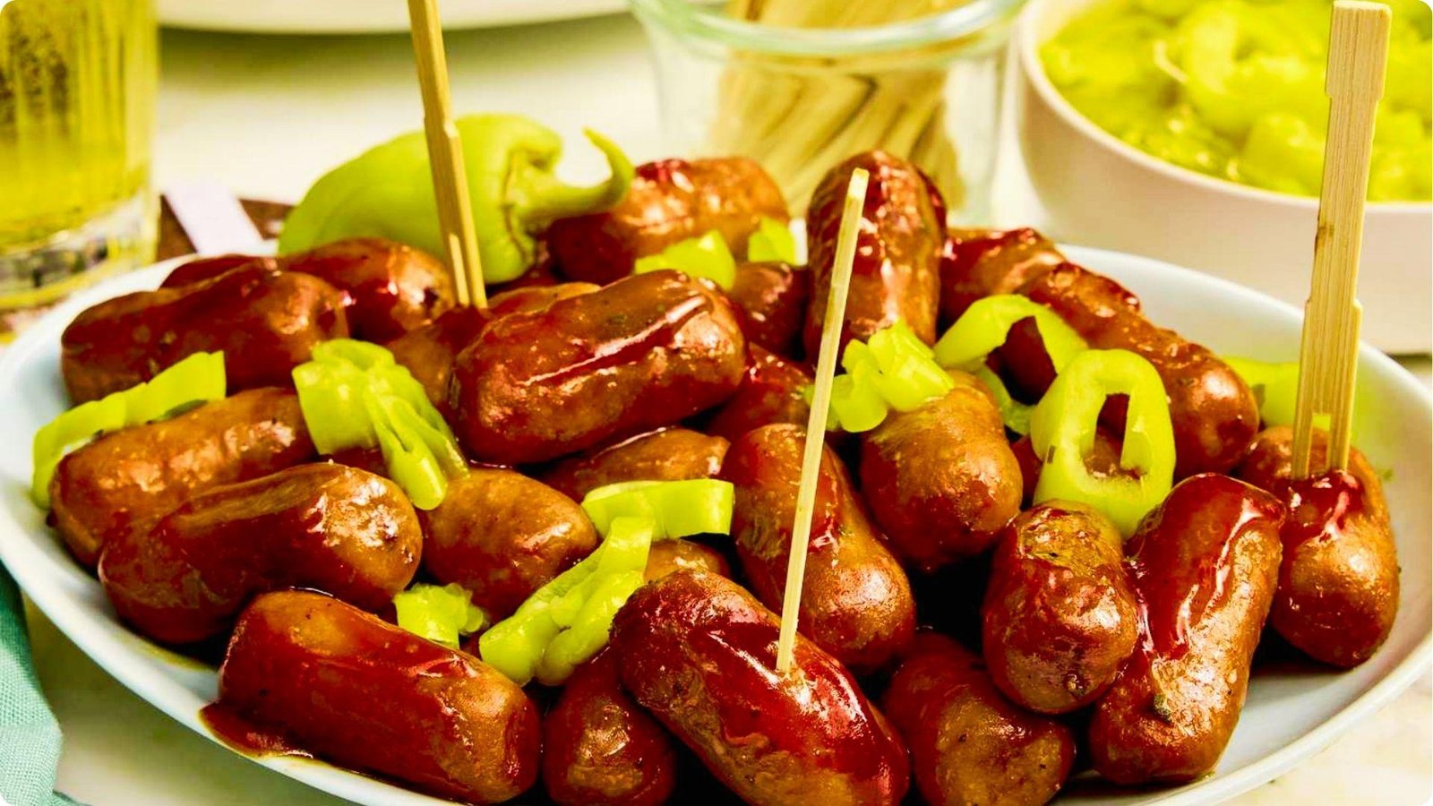 A large plate of BBQ-glazed cocktail wiener with green jalapeno slices on toothpicks, served with guacamole dip.