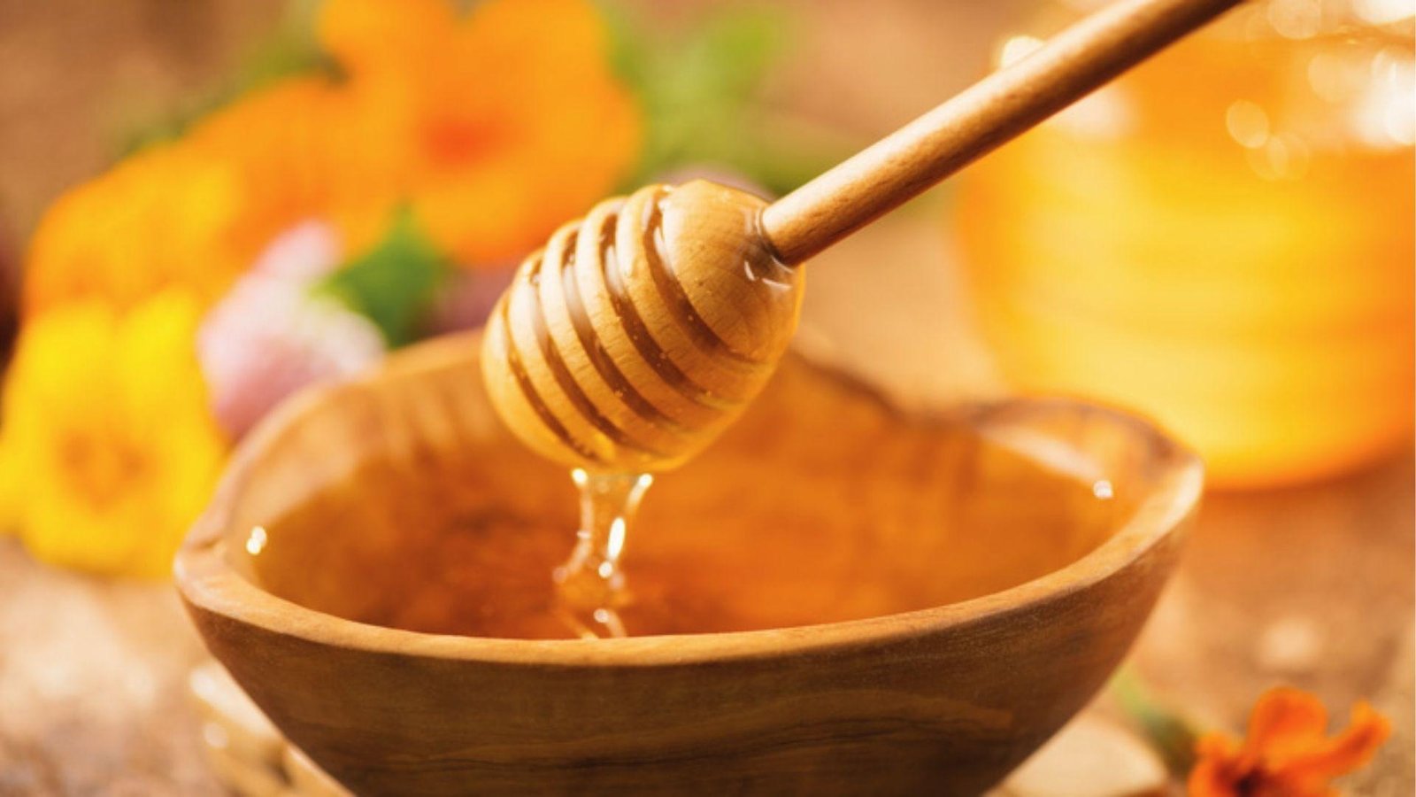 Honey dipper lifts rich honey from a wooden bowl with vibrant marigold flowers in the background.