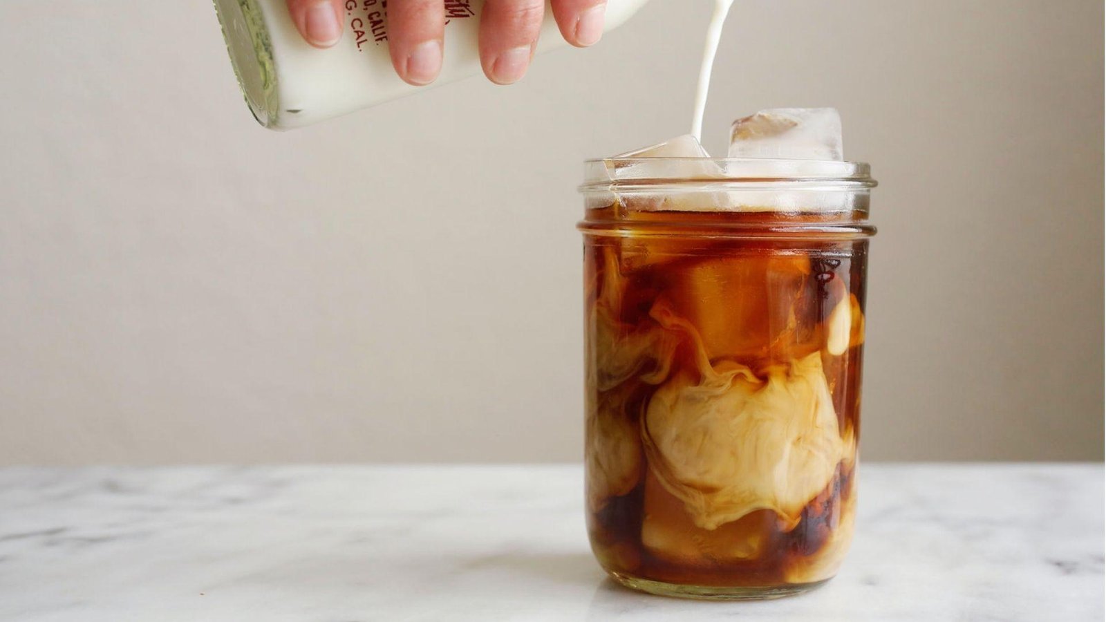 A hand pouring cream from a glass bottle into a mason jar of iced black coffee on a marble surface.