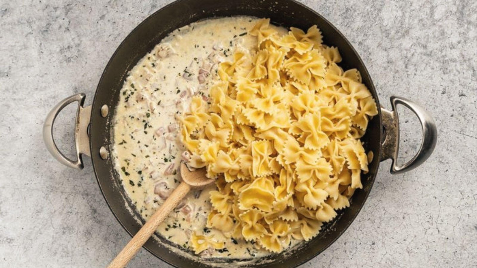 Farfalle pasta added to creamy herb sauce with bacon bits in black pan, overhead wooden spoon