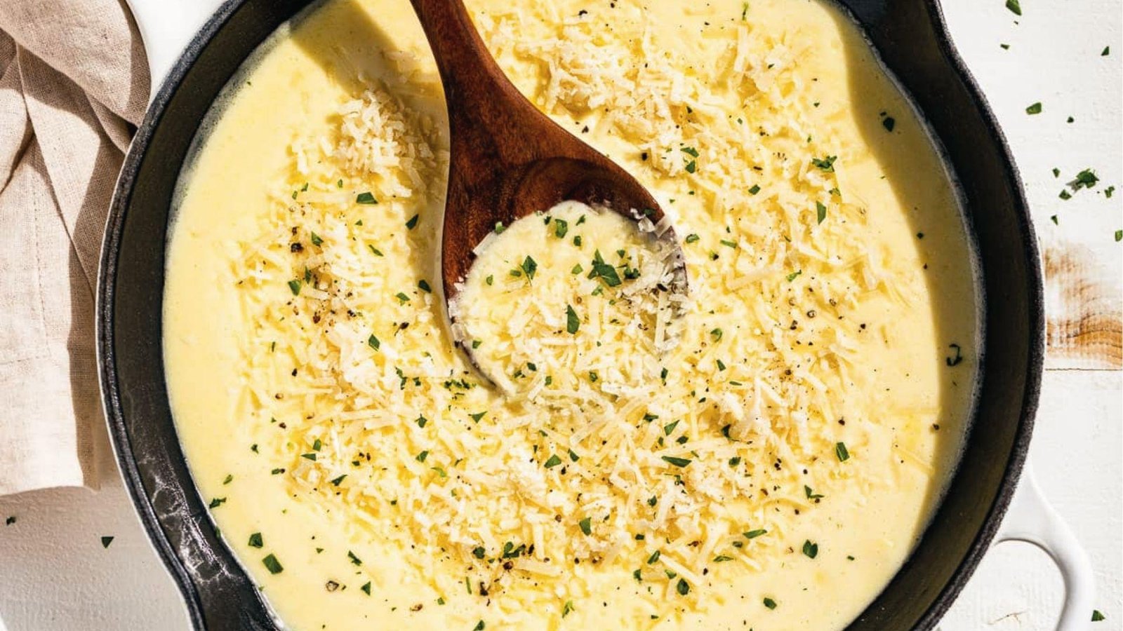 Rich yellow cheese cream sauce with herbs and black pepper in cast iron skillet, wooden spoon
