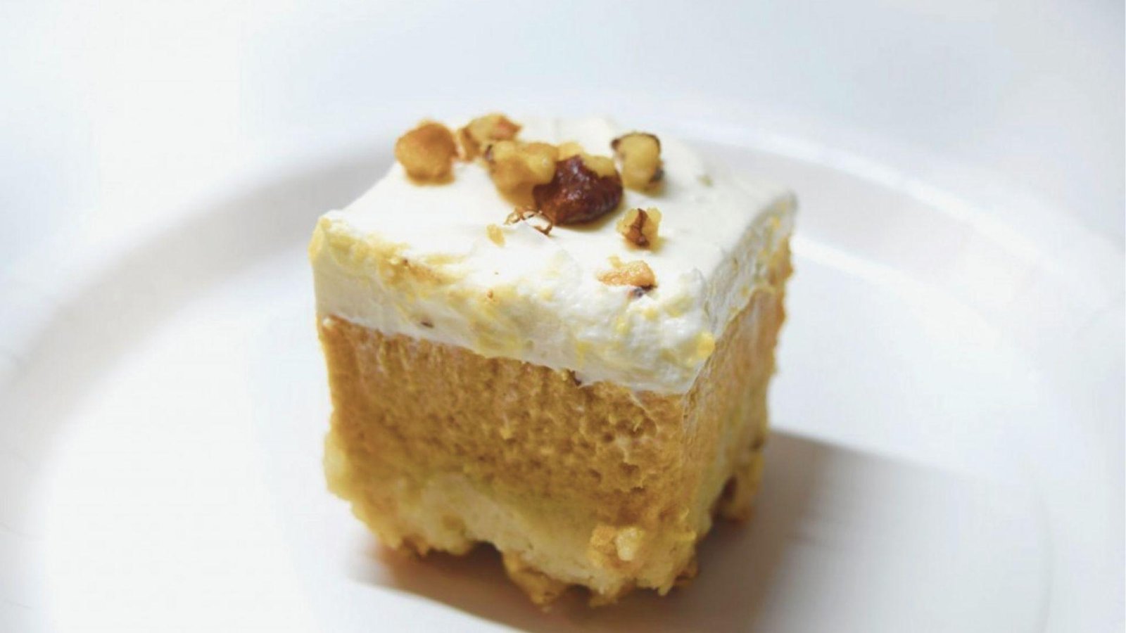Close-up of a Pumpkin Crunch dessert square on a white plate with whipped cream frosting and walnut pieces on top.