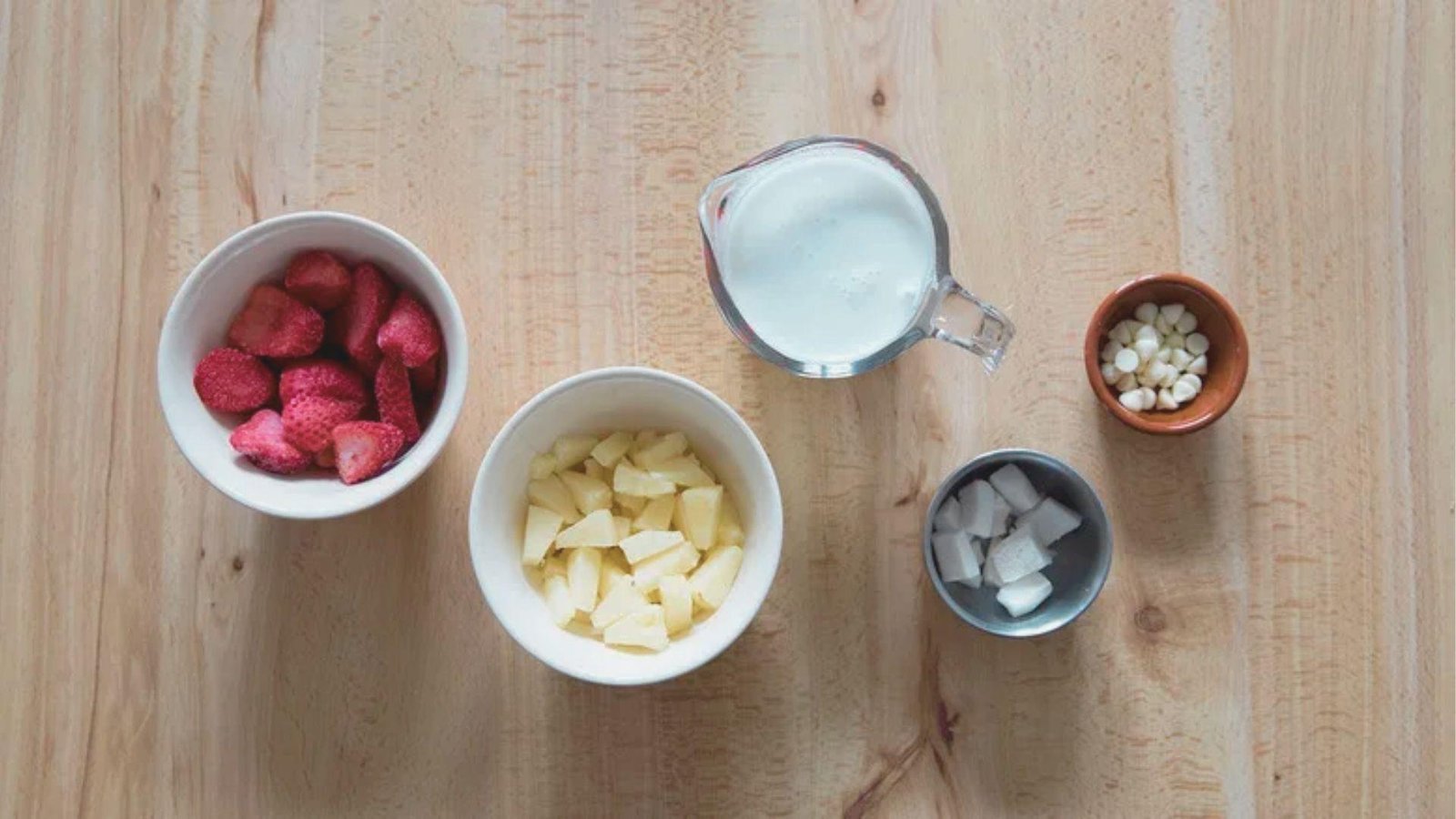 Flat lay of Bahama Mama Tropical Smoothie ingredients on a wooden table: frozen strawberries, pineapple chunks, coconut milk, coconut chunks, and white chocolate chips.