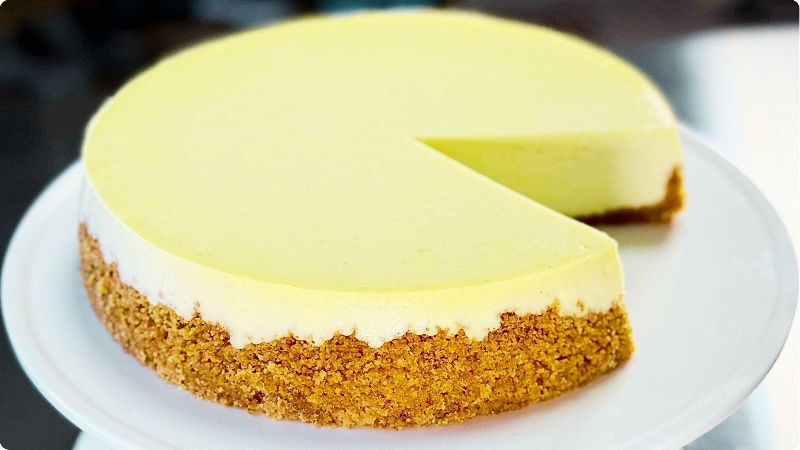 A fully baked 6-inch cheesecake with a smooth, pale yellow top rests on a white cake stand with one slice cut out.