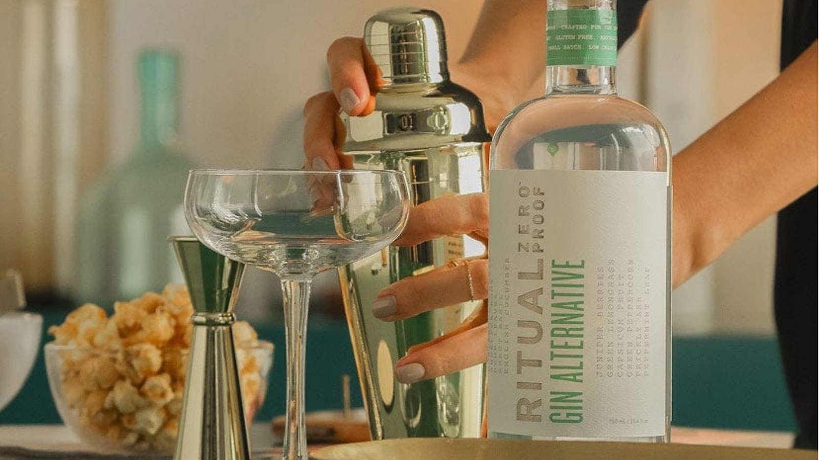 A woman's hand holding a cocktail shaker beside a coupe glass and a bottle of Ritual Zero Proof Gin Alternative.