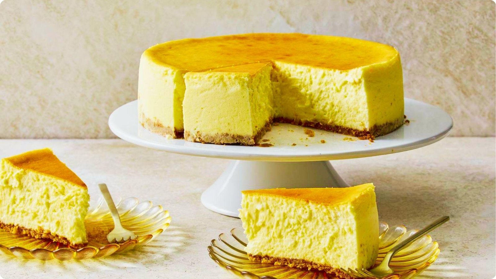 A golden-baked 6-inch cheesecake sits on a white cake stand with one slice cut and two slices plated on scalloped glass dishes.