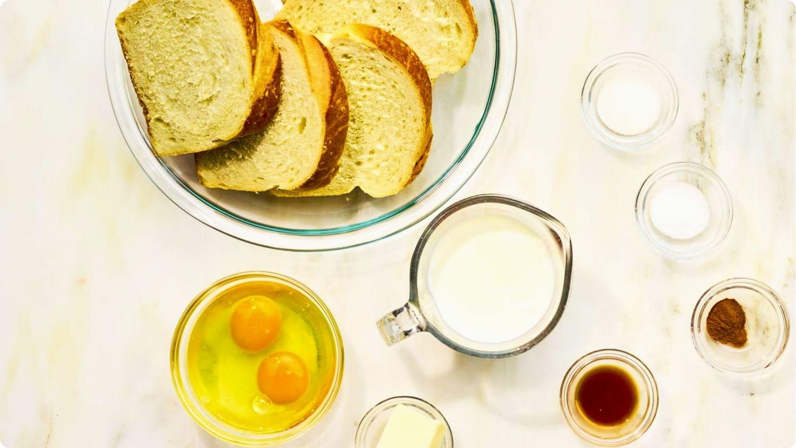 Wheat bread slices, eggs, milk, cinnamon, vanilla, sugar, and butter arranged on a marble counter.