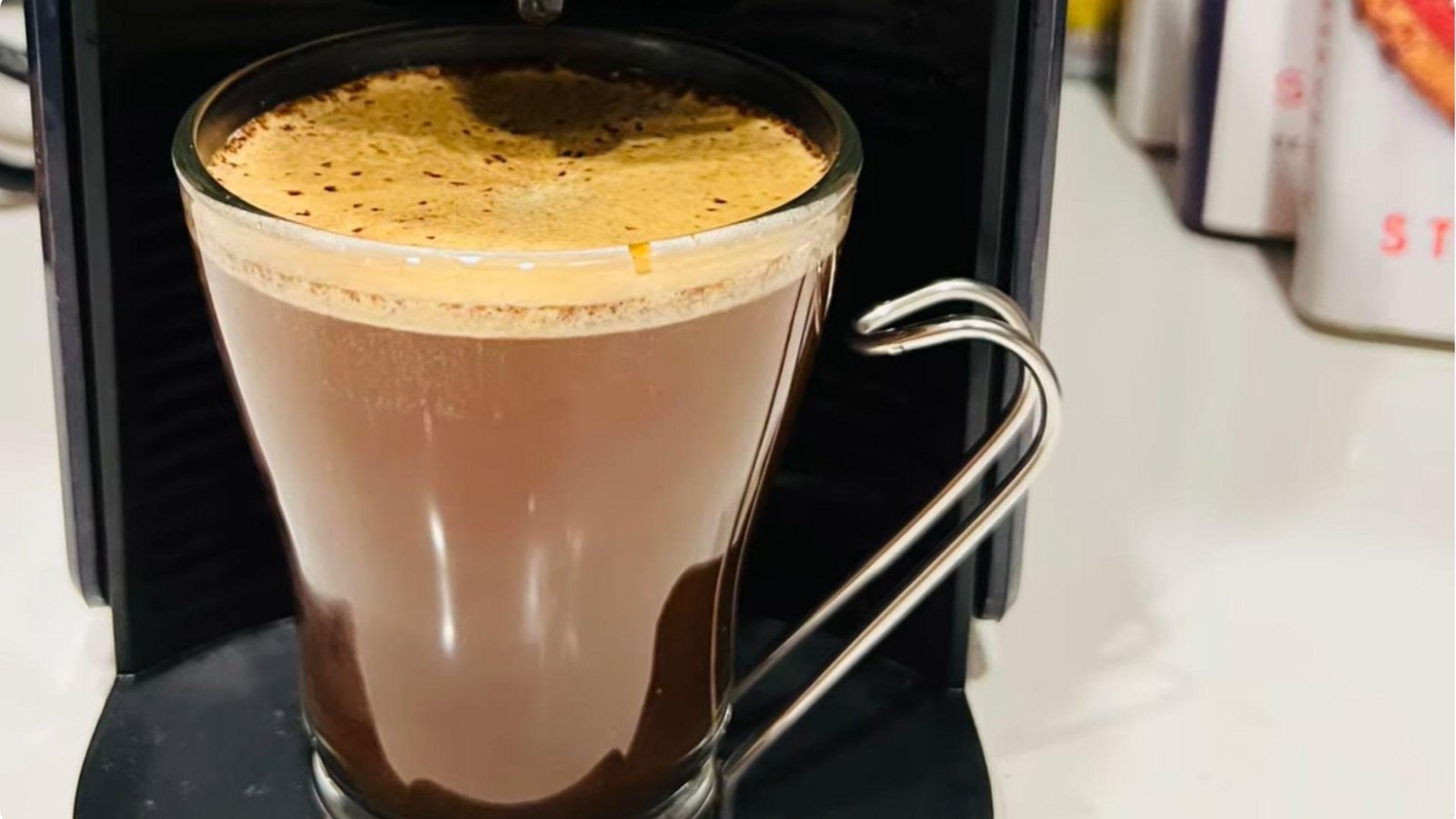 A clear glass mug with a metal handle filled with frothy mushroom coffee sitting in front of a pod coffee machine.