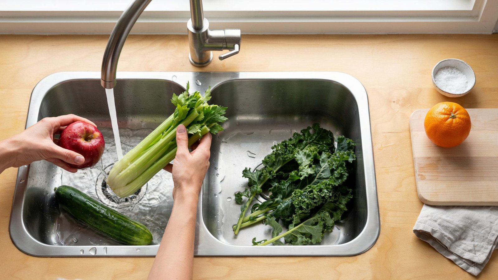 Whole apple celery cucumber and kale being rinsed under water with orange kept aside on dry counter near cutting board.