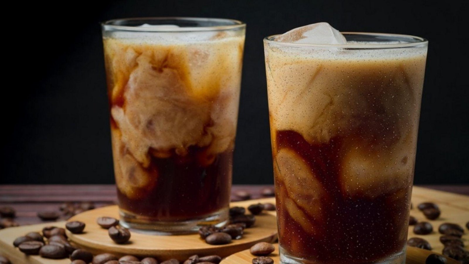 Two glasses of iced coffee with swirling cream and ice cubes surrounded by scattered coffee beans on a wooden board.