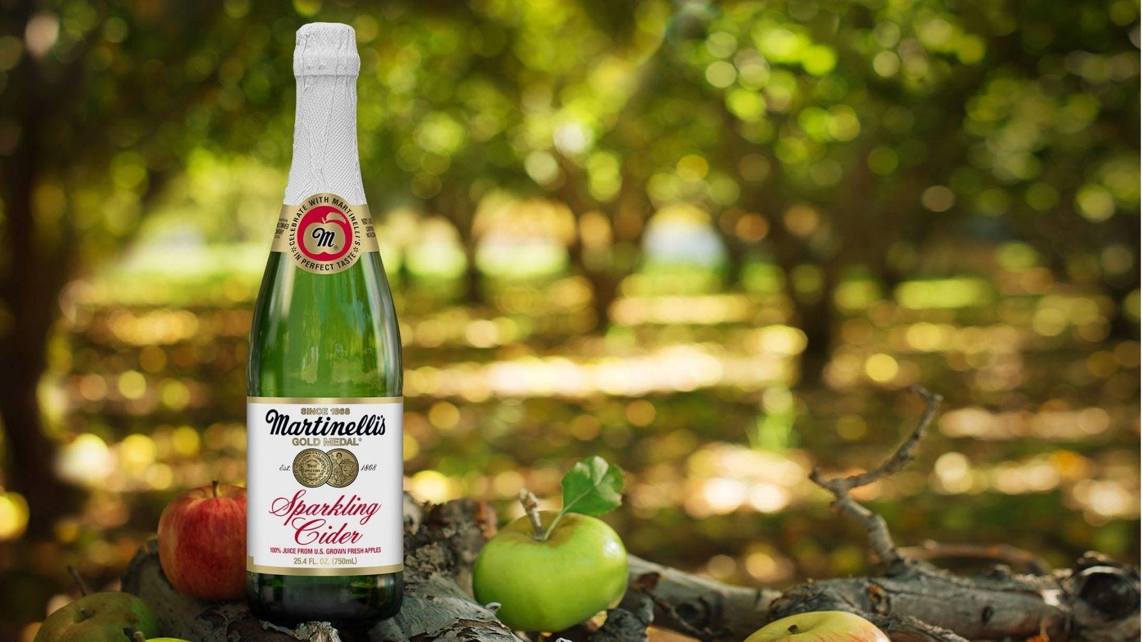A bottle of Martinelli's Gold Medal Sparkling Cider standing among red and green apples in a sunlit orchard setting.