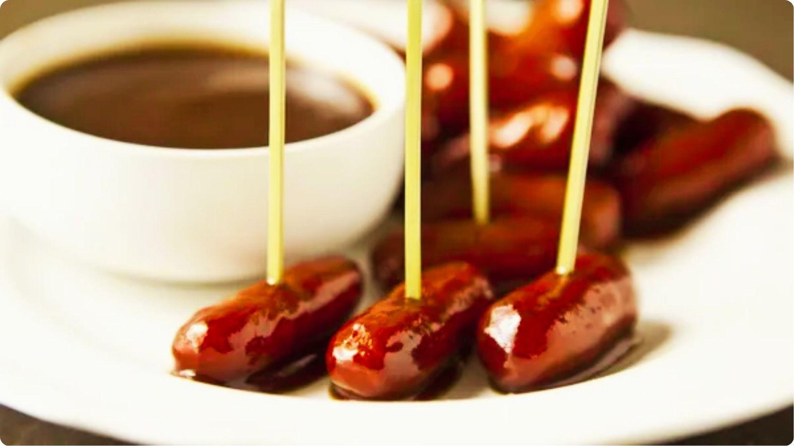 Glazed mini sausages on toothpicks arranged on a white plate with a dark dipping sauce in a small bowl.