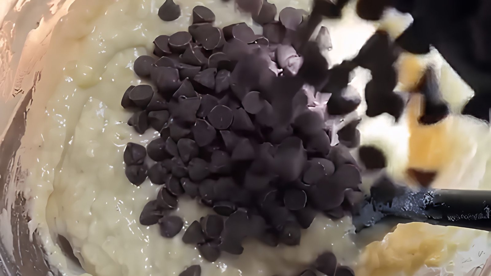 Chocolate chips being gently folded into banana muffin batter to evenly distribute sweet chocolate pieces throughout.