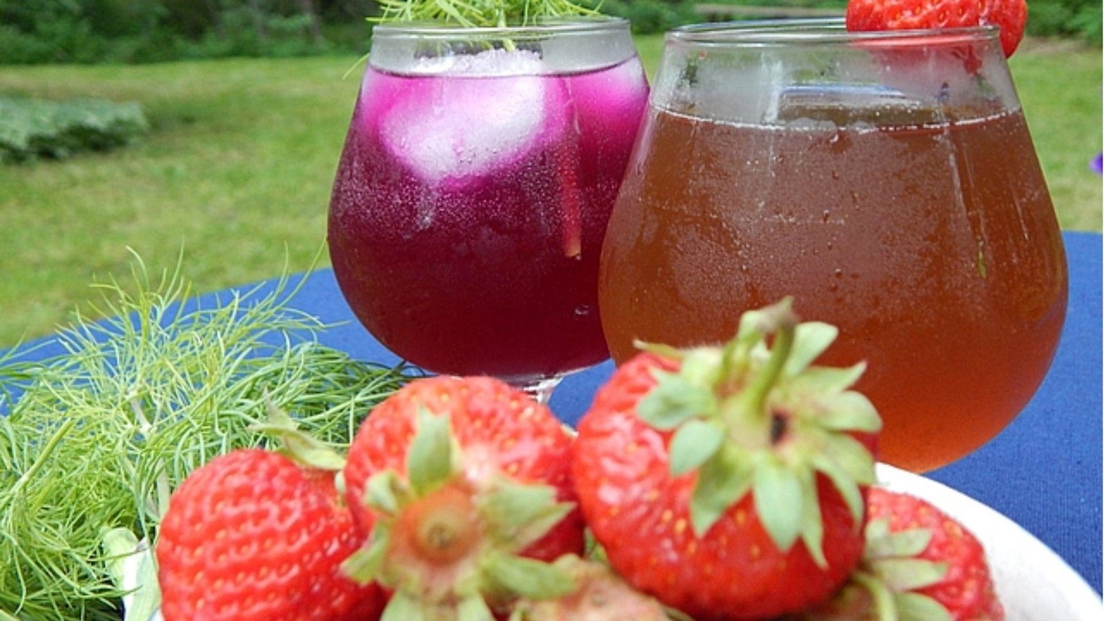 Two bubble glasses of purple and amber iced drinks with a bowl of fresh red strawberries on a blue outdoor table.