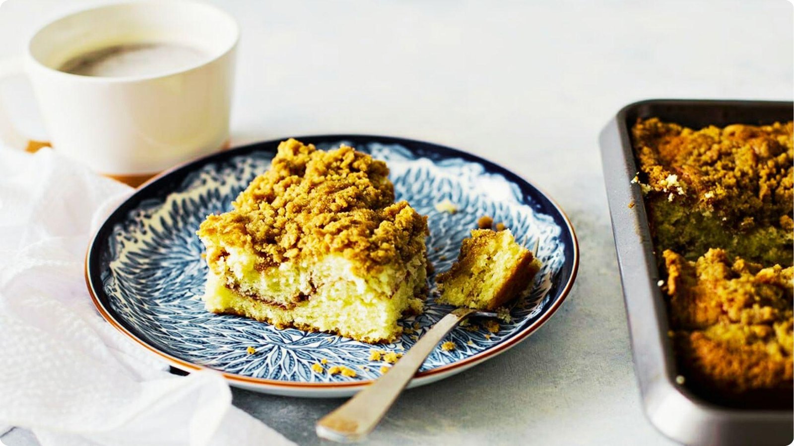A slice of German coffee cake with golden streusel topping on a blue patterned plate, next to a baking pan.