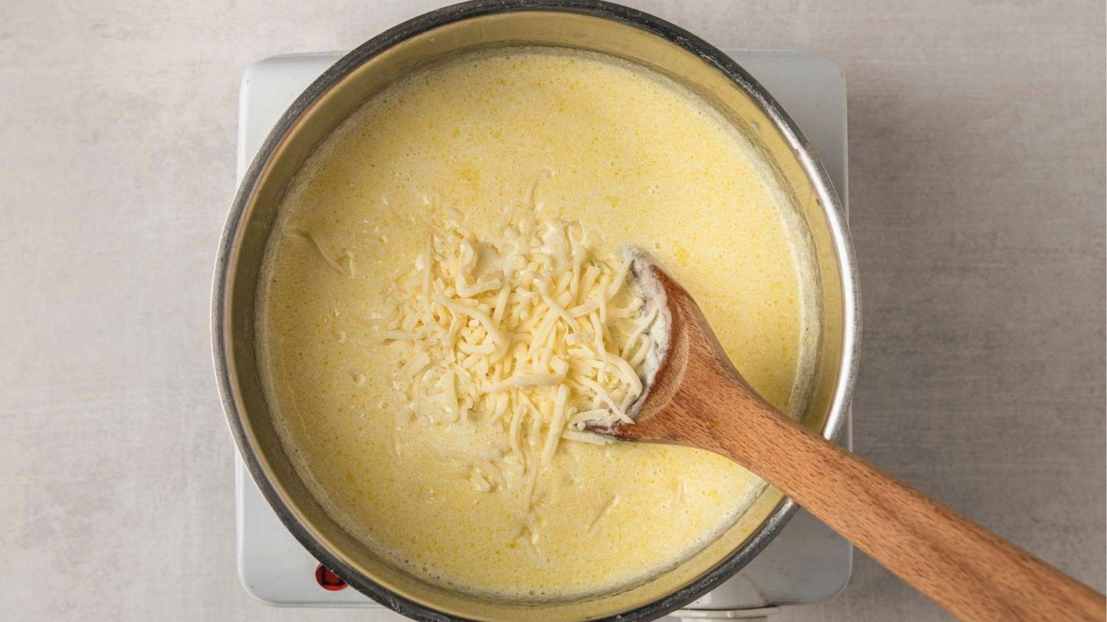 Parmesan cheese melting into yellow cream sauce in saucepan with wooden spoon on hot plate