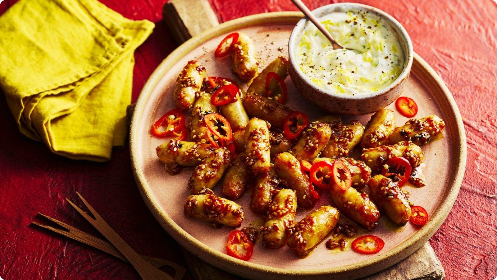Glazed mini wieners topped with sesame seeds and sliced red chilli on a plate with a creamy herb dipping sauce.