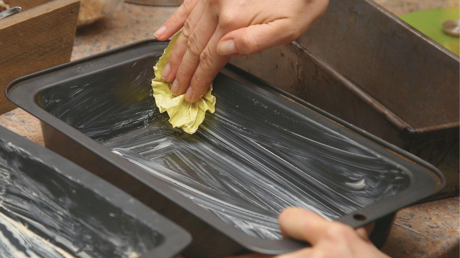 Hand greasing a dark loaf pan with butter using a paper towel to prevent sticking.