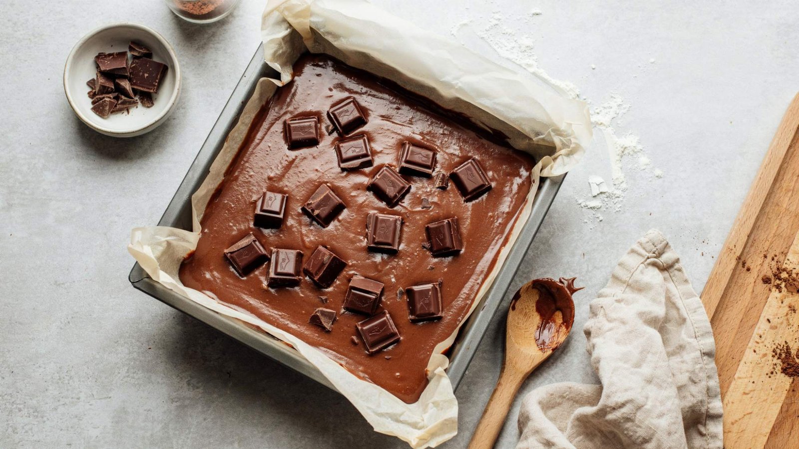 Brownie batter spread in a parchment-lined baking pan topped with dark chocolate chunks, ready to bake.