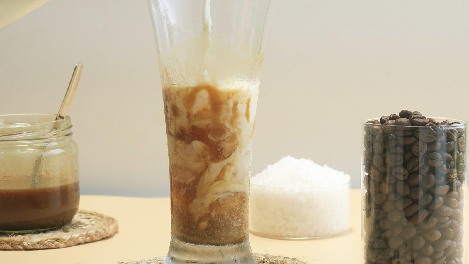 Milk being poured into a tall iced caramel coffee glass beside a jar of espresso, sugar, and coffee beans.